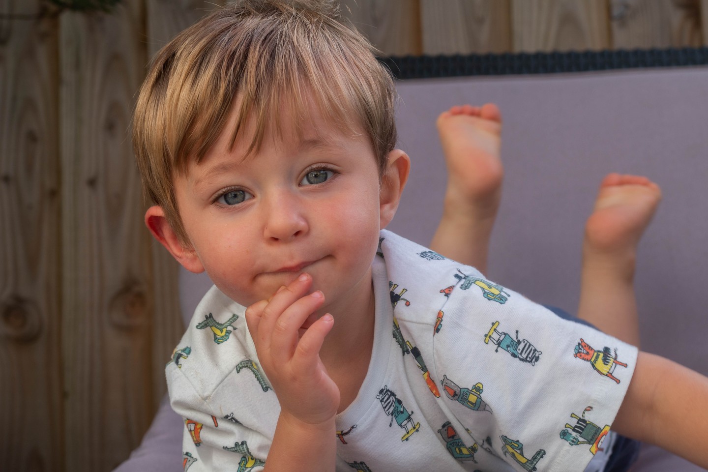 A young boy with light brown hair and blue eyes lies on his stomach, feet raised in the air, hand near his mouth, wearing a patterned t-shirt.