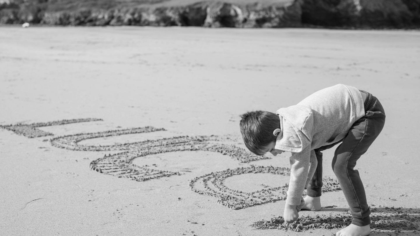 Black and white photo of a barefoot boy bending over to write large letters in sand on a beach, with rocky cliffs in the background.