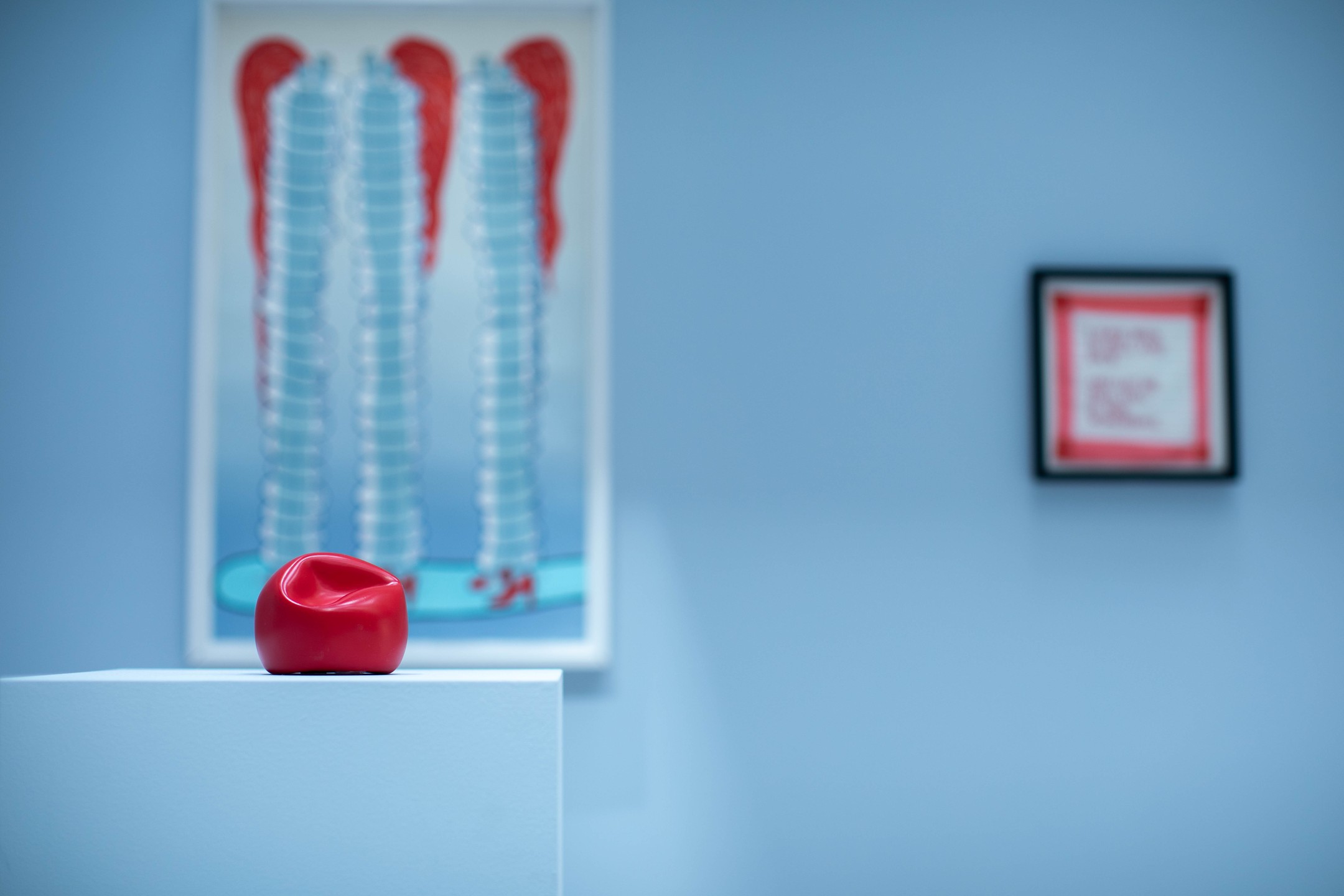 Gallery scene: small red glossy sculpture on white pedestal, framed print of three ribbed towers with red tops, and distant framed red-bordered artwork on blue wall.