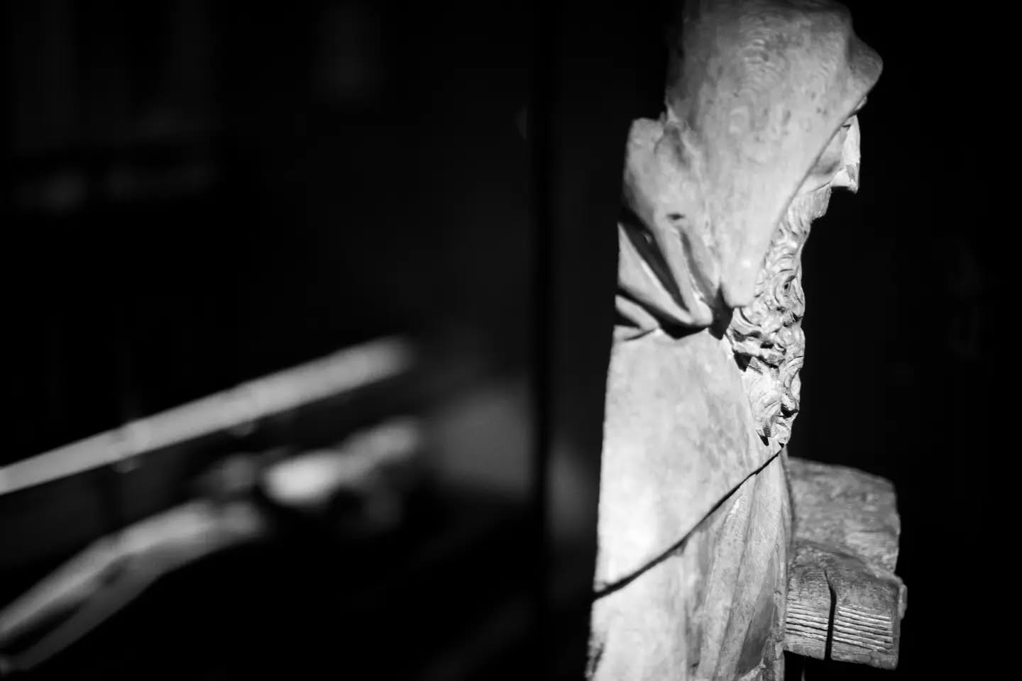Black-and-white photo of a weathered stone statue's draped hood and shoulder, lit against a dark blurred interior background.