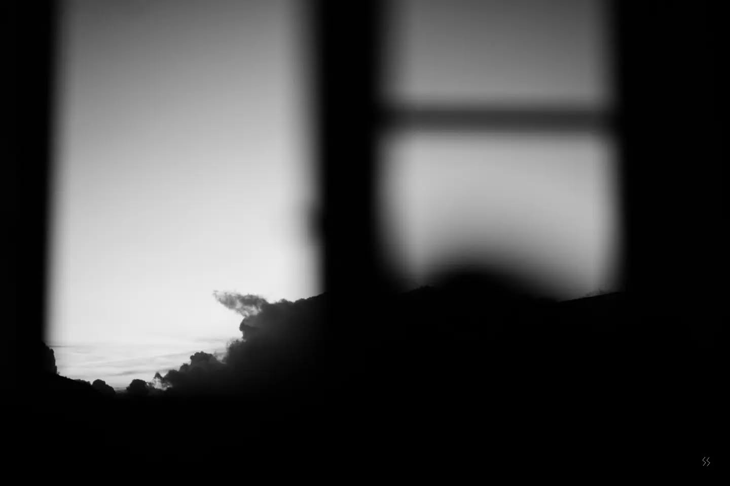 Black and white blurred image of two window frames, one showing pale sky and distant hazy hills, the other softly blurred glass panes.