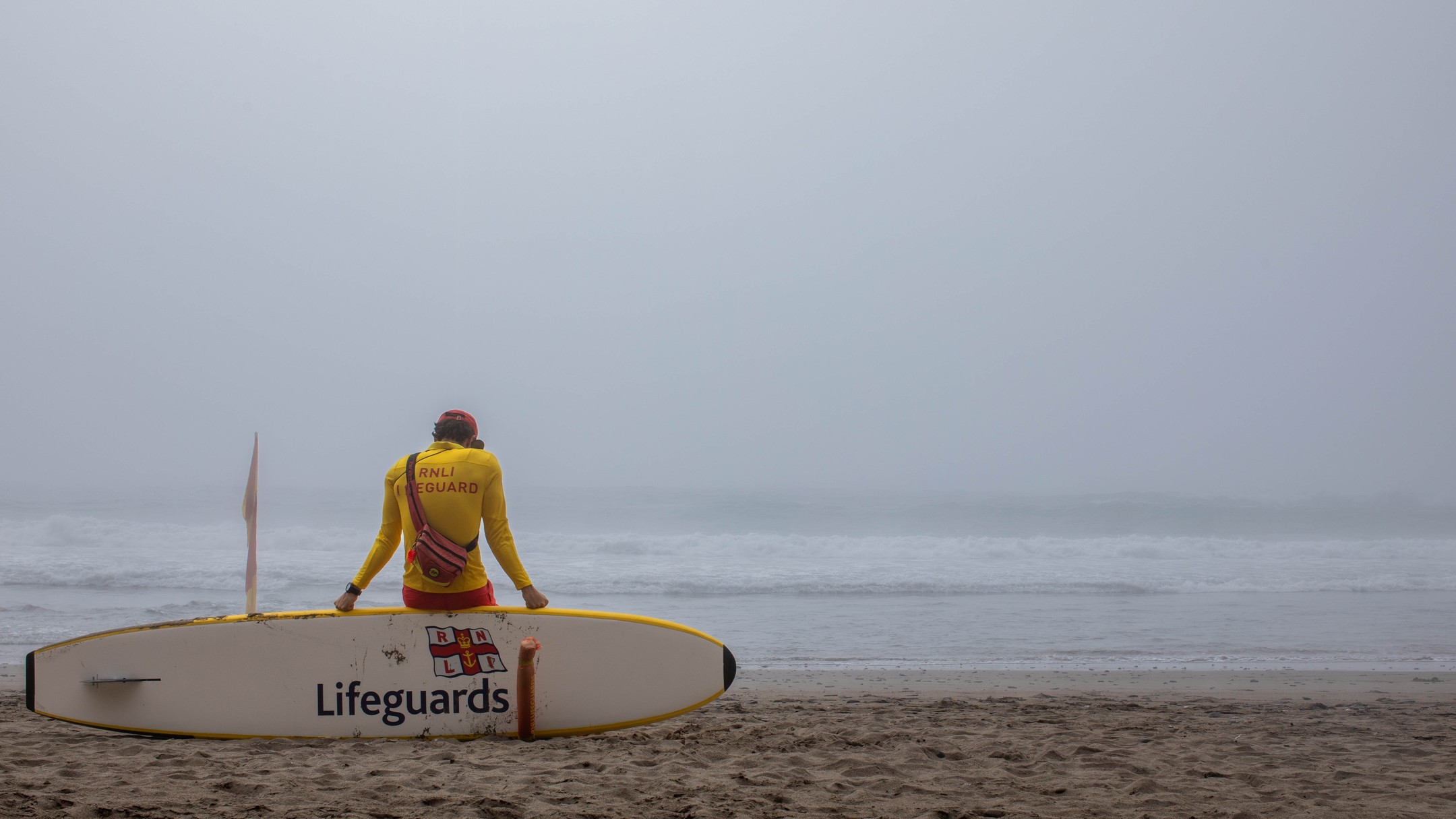 A lifeguard in a yellow and red uniform sits on a rescue surfboard on a beach, facing a foggy grey sea, with a flag beside them.