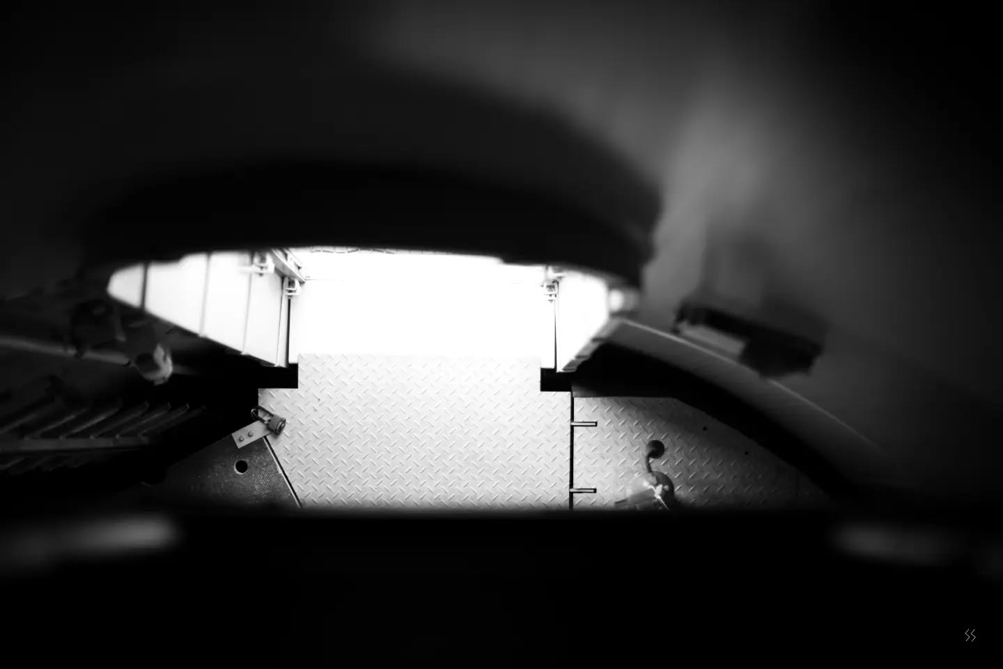 Black and white photo through a narrow gap showing a bright diamond-plate metal floor and industrial machinery framed by dark blurred edges.