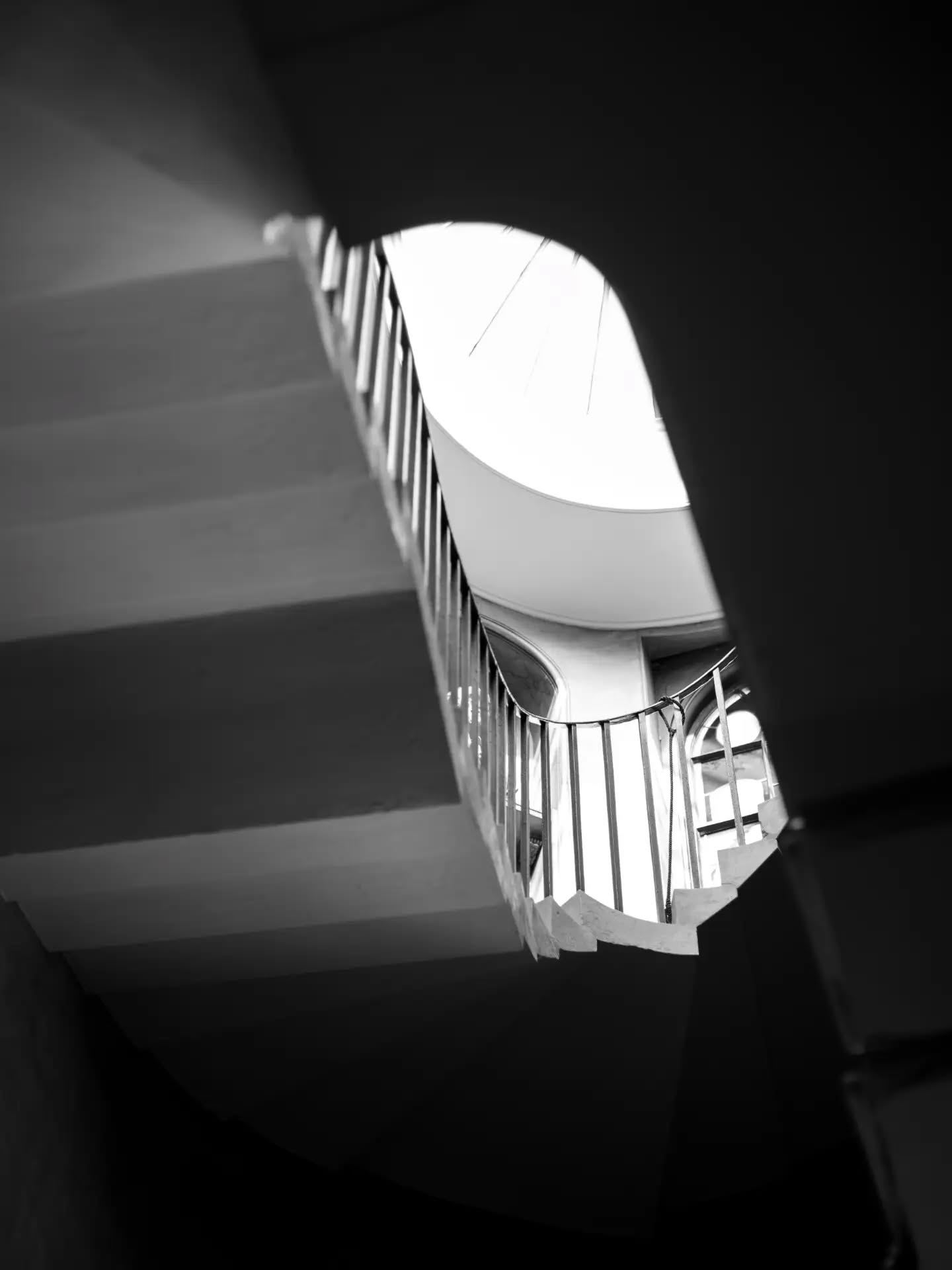 Black and white photo of a spiral staircase with metal railing, viewed from below looking up toward a bright skylight.