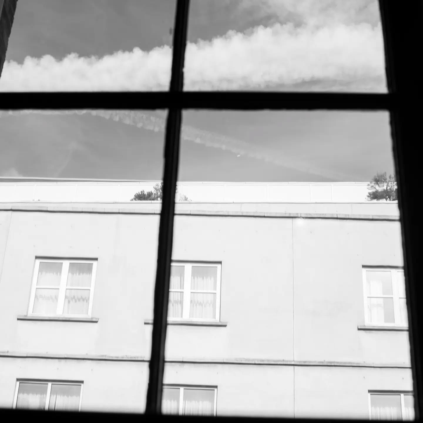 Black and white photo through window panes showing a building facade with windows and curtains, sky with contrails above.