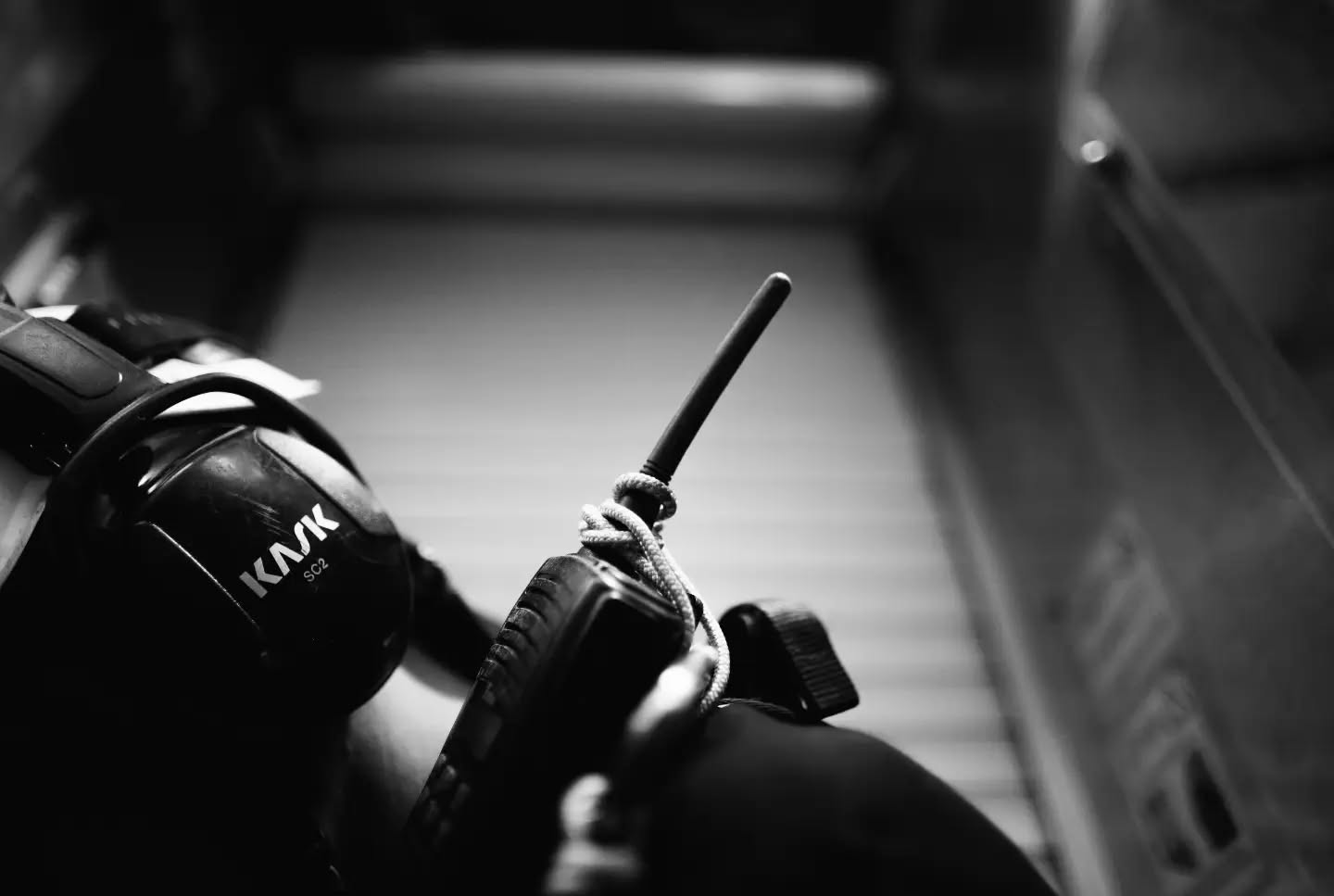 Black and white close-up of a hand holding a two-way radio with antenna, next to a Kask helmet, in a corridor.