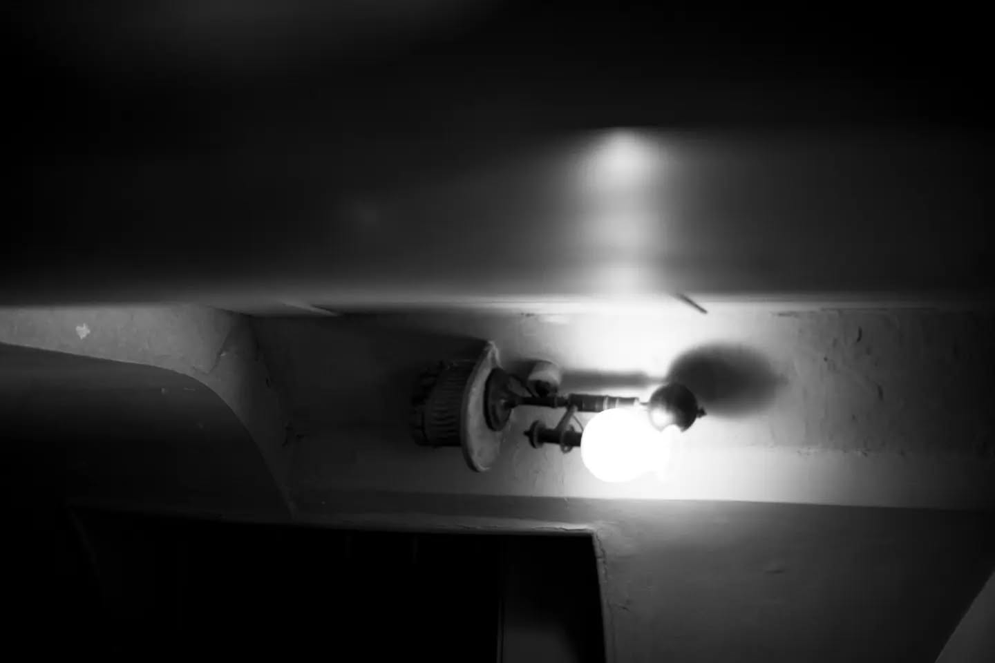 Black-and-white photo of a bare bulb mounted on a ceiling fixture, glowing against dark surroundings and casting shadow.
