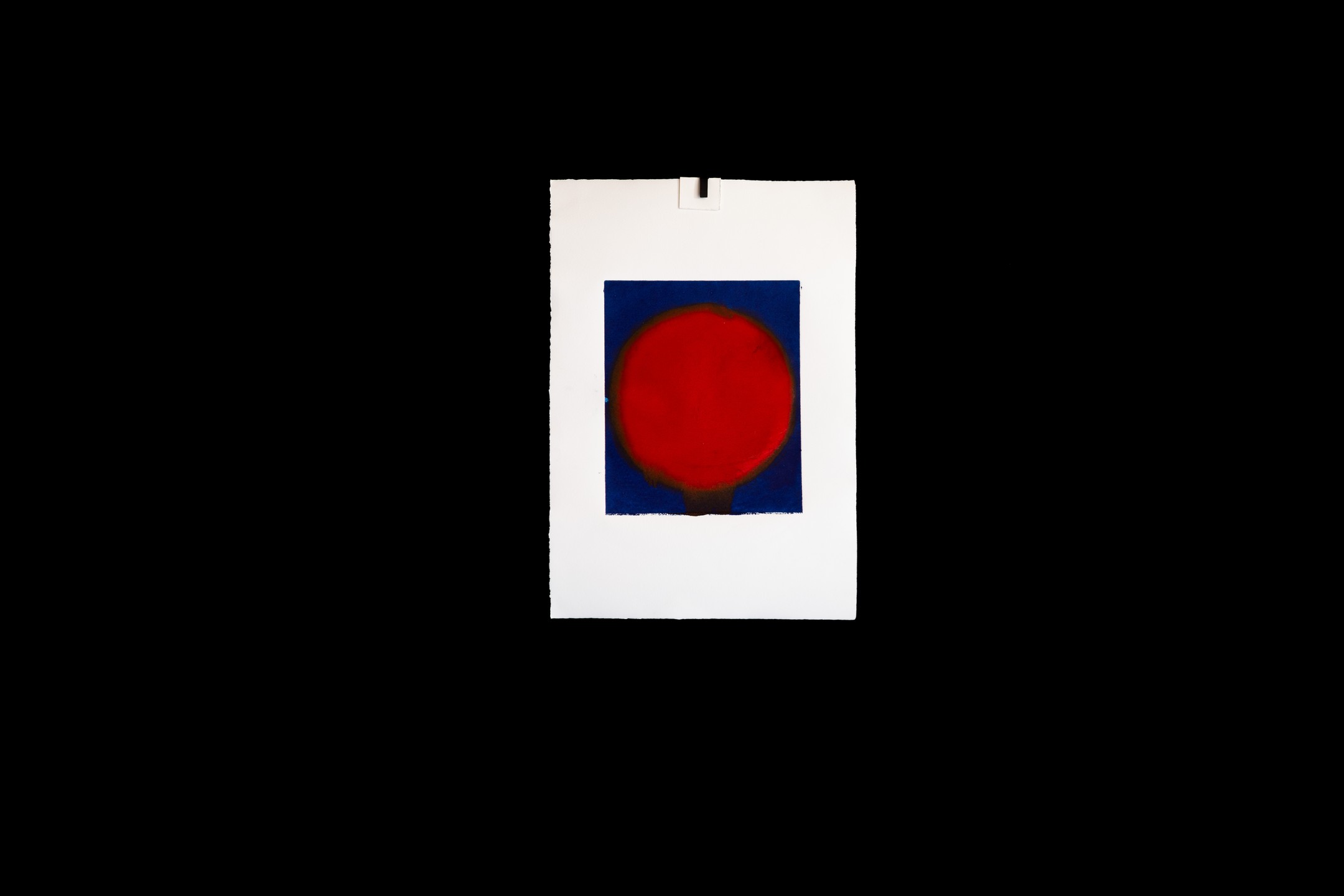 A print on white paper showing a red circular form on a dark blue rectangular background, mounted against a black backdrop.