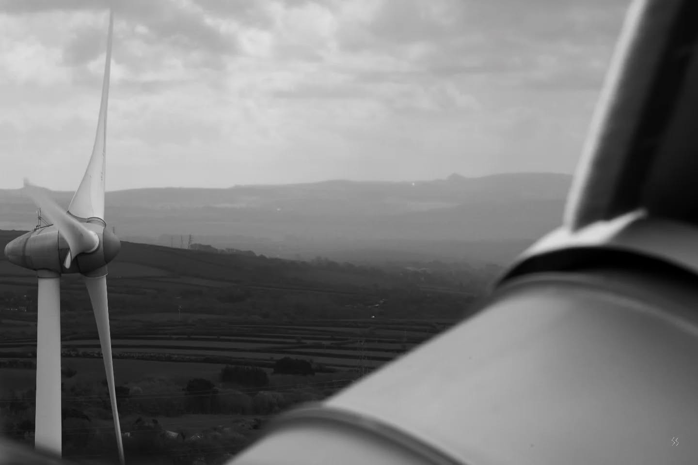 Black and white image of a wind turbine's blurred blade in foreground, with a second turbine standing on a hillside amid patchwork fields and hazy hills beyond.