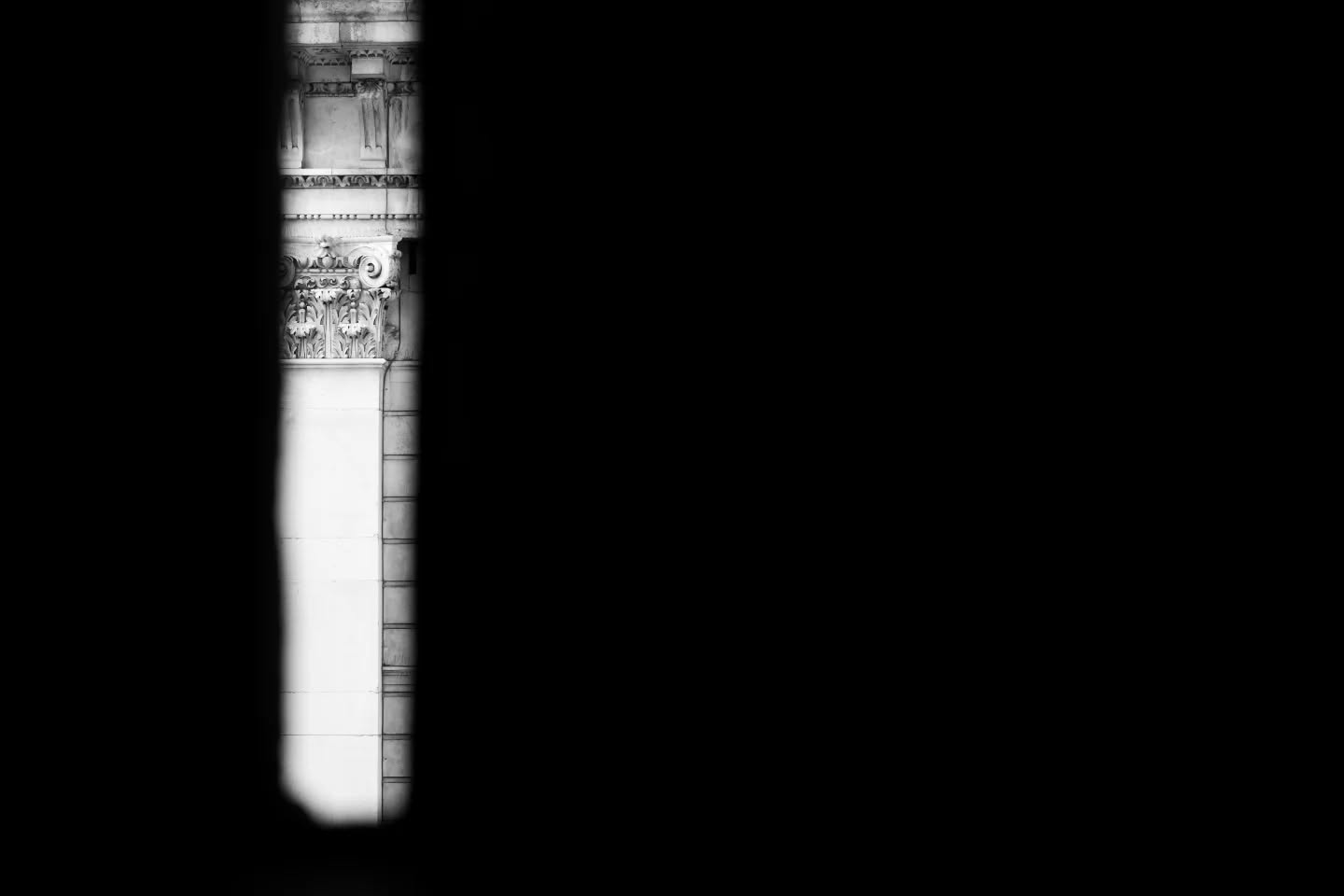 A narrow sliver of light reveals a classical stone column with an Ionic-style capital, flanked by deep black shadow on either side.