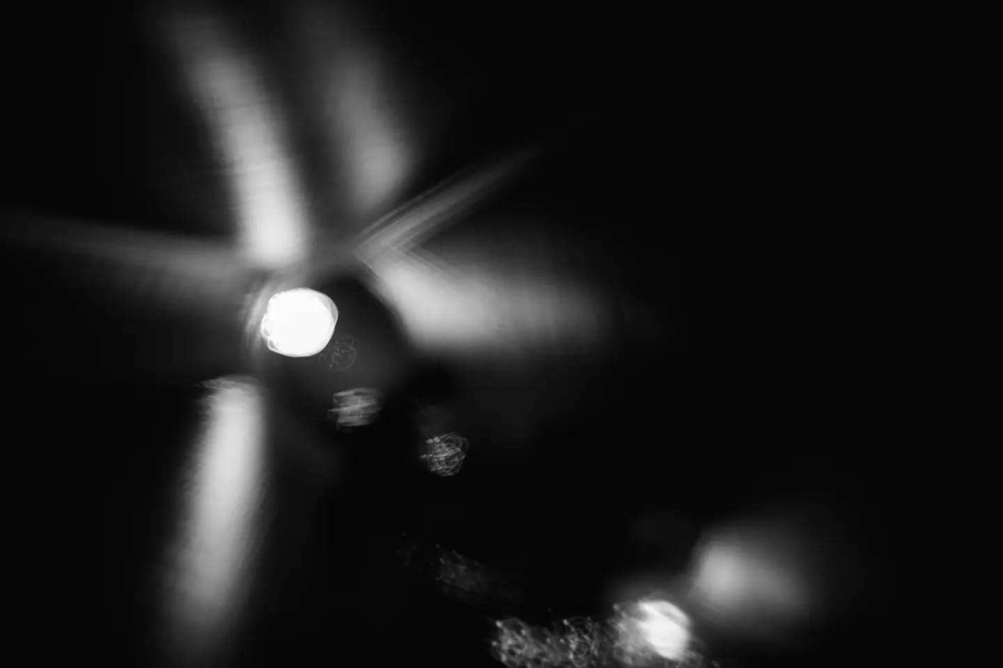 Black and white abstract long-exposure image of a bright light source with radiating streaks, surrounded by darkness and smaller blurred light spots.